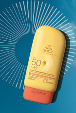 Protetor Solar Infantil FPS 50 Natura Solar em fundo de céu azul com nuvens e grafismos brancos. Embalagem amarela com tampa laranja e detalhes em preto.