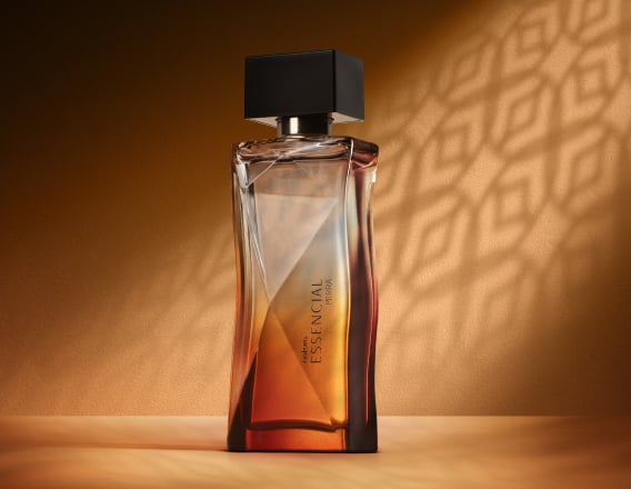 Deo Parfum Essencial Mirra Feminino em frasco facetado degradê âmbar com tampa preta. Está em fundo cor areia com sombra geométrica projetada.