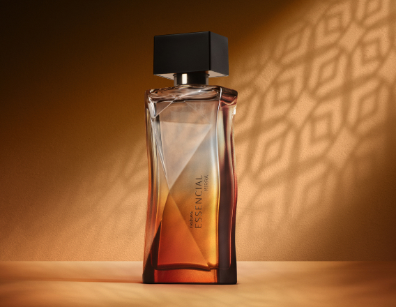 Deo Parfum Essencial Mirra Feminino em frasco facetado degradê âmbar com tampa preta. Está em fundo cor areia com sombra geométrica projetada.