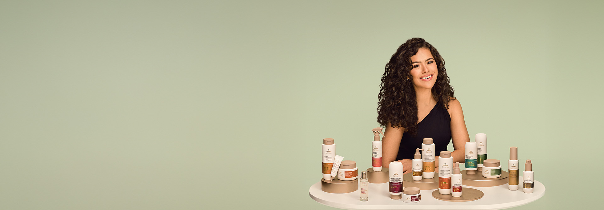 Maisa sorrindo ao lado da linha completa Natura Lumina, disposta sobre uma mesa branca com fundo verde.