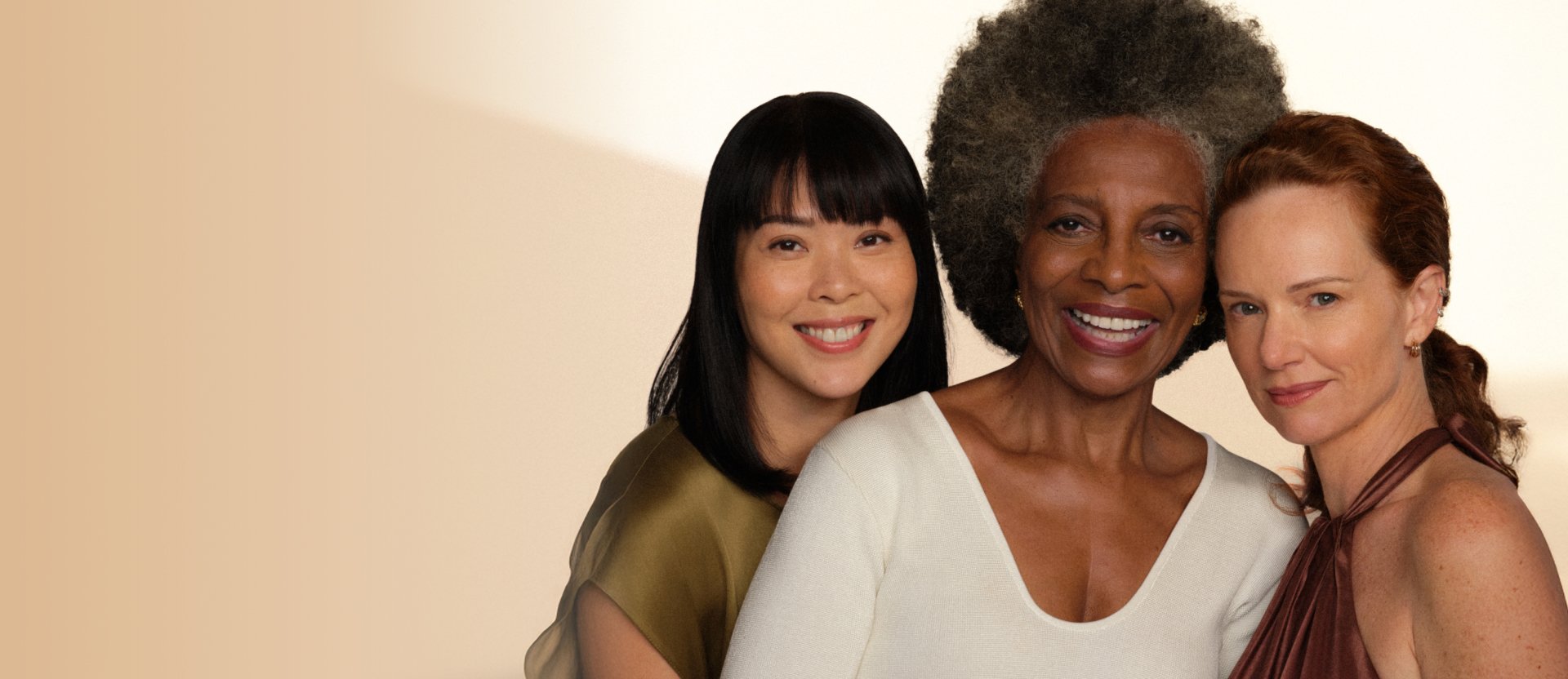 Três mulheres de diferentes faixas etárias e etnias sorriem para a câmera. Elas usam roupas em tons terrosos, em fundo claro.
