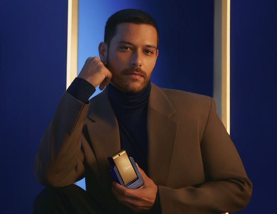 Homem de cabelos curtos, usando camisa de gola alta preta e terno marrom, segura a fragrância Natura Homem Evolut.io próximo ao corpo, em fundo azul e moldura branca iluminada.