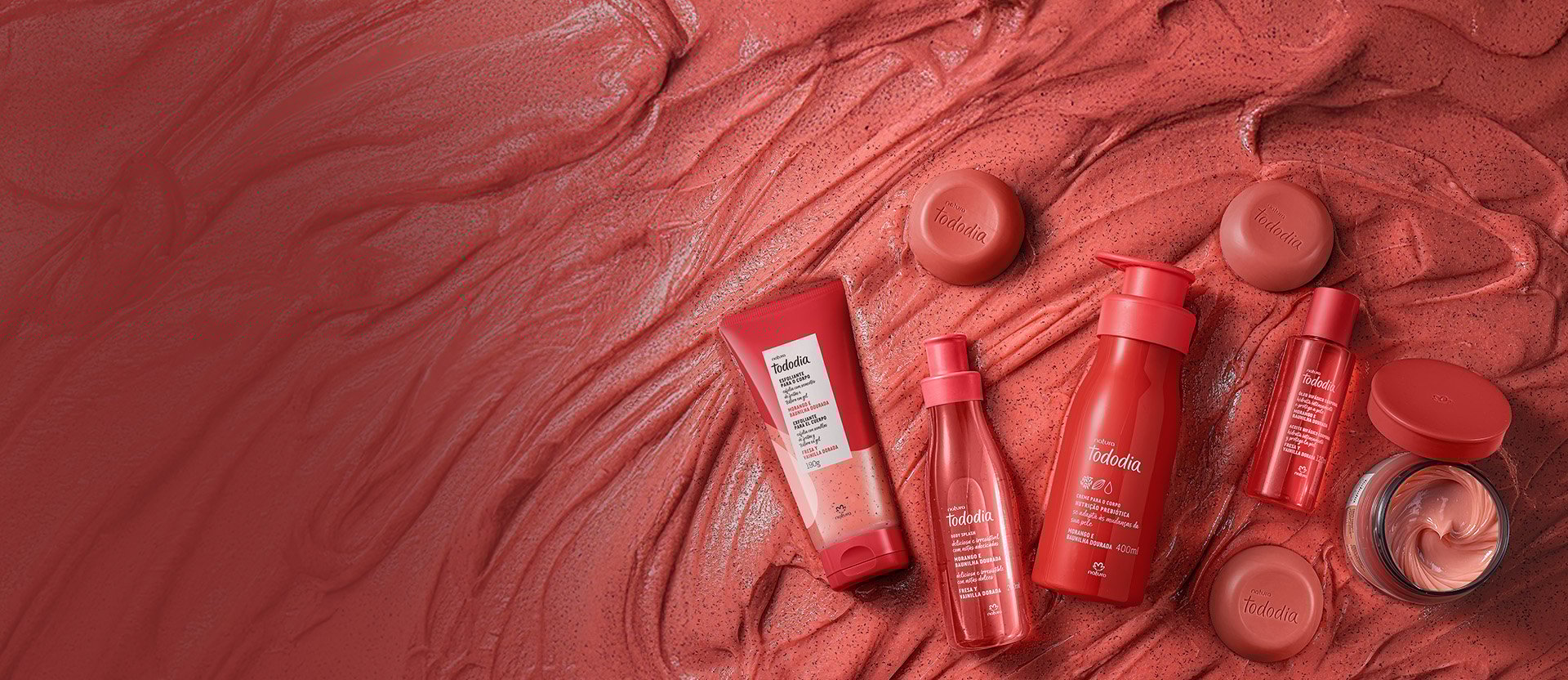 Produtos da linha Natura Tododia Morango e Baunilha Dourada dispostos sobre uma textura cremosa de cor rosa. A imagem inclui creme para o corpo, body splash, esfoliante, sabonetes em barra e um pote de creme aberto, todos em embalagens de tons avermelhados.