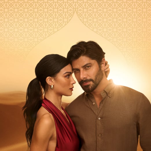 Uma mulher branca de cabelo escuro preso e um homem branco com cabelo castanho e barba volumosa se abraçam, com os rostos próximos. Fundo areia, com padrões geométricos e feixe de luz.