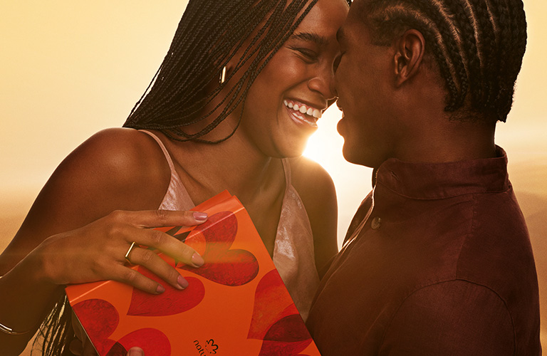 Foto de um casal se beijando ao pôr do sol, com céu alaranjado ao fundo, enquanto a mulher segura uma caixa laranja com estampa de corações.