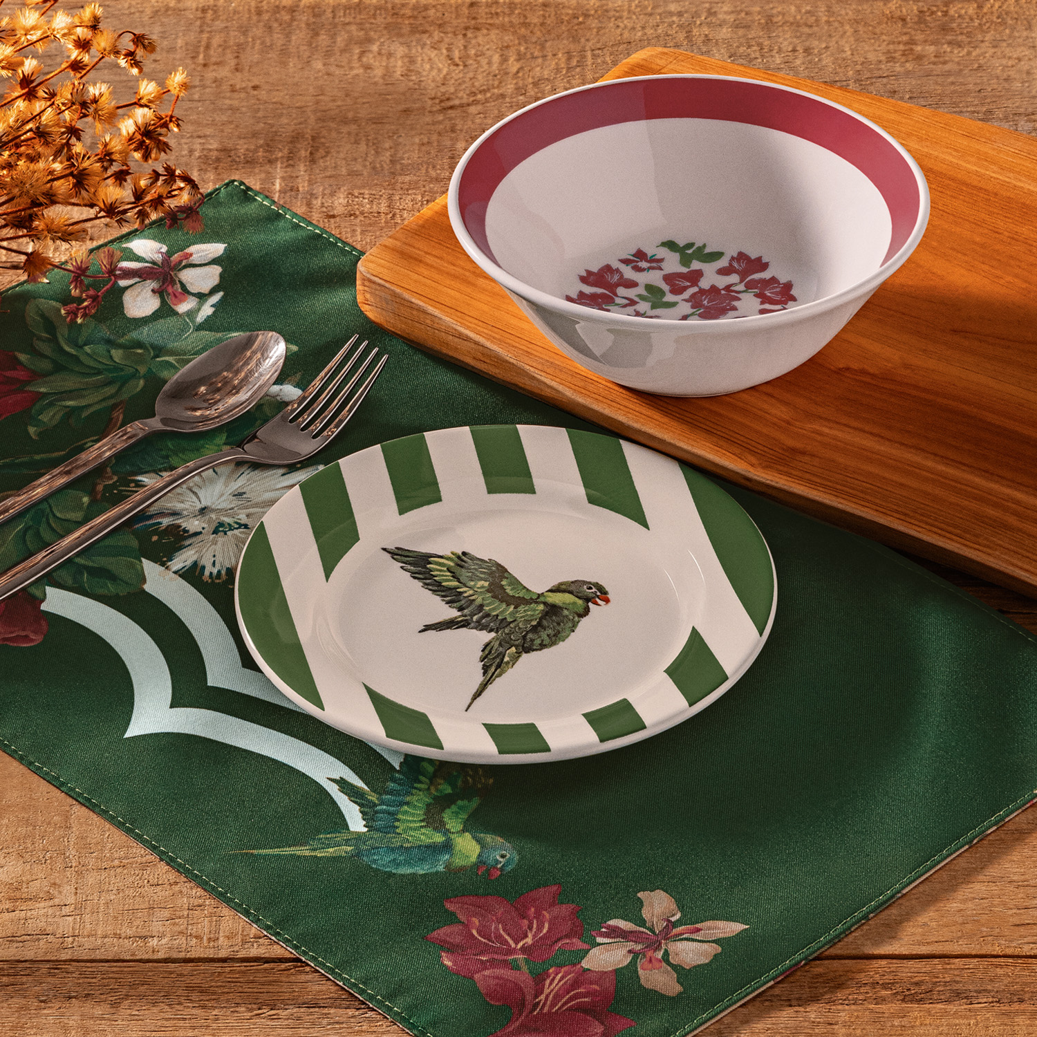 Mesa de madeira montada com 3 produtos de Crer Para Ver: jogo americano, prato de sobremesa e tigela de cerâmica. Itens em tons de verde escuro, branco e vermelho com ilustração de pássaro e flores. 