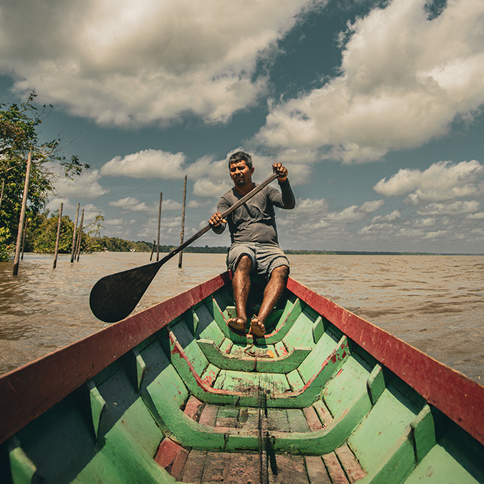 Foto de homem ribeirinho em embarcação e remo nas mãos em rio amazônico.