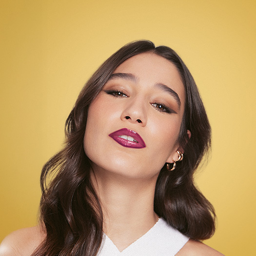 Mulher branca de cabelo castanho-escuro usa maquiagem marcante, incluindo batom roxo brilhante e delineador. Ela usa brincos de argola dourados e uma blusa branca, diante de fundo amarelo vibrante.