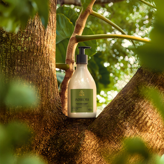 Frasco de creme com válvula preta de Polpa Desodorante Hidratante para o Corpo Ekos Andiroba sobre tronco de árvore, em cenário de floresta desfocada.