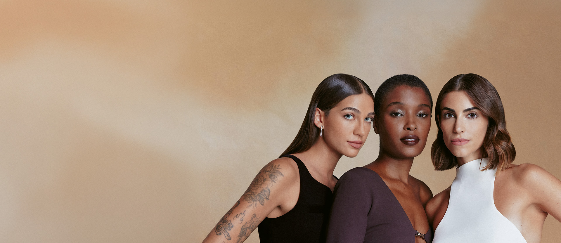Três mulheres posam lado a lado sobre um fundo bege-claro. Uma mulher branca com cabelo escuro e tatuagens, uma mulher negra de cabelo curto e uma mulher branca com cabelo castanho.