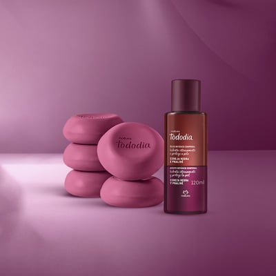 Linha Tododia Cereja Negra e Praliné com 5 sabonetes redondos, 1 frasco de body splash, 1 pump de creme, 1 pote de creme veludo e 1 frasco de óleo corporal. Embalagens roxas em fundo roxo iluminado.