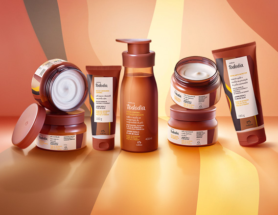 Em fundo em tons de marrom e amarelo, produtos da linha Natura Tododia Jambo Rosa e Flor de Caju: Óleo em Creme, Geleia Desodorante, Loção Corporal, Manteiga Desodorante e Esfoliante Nutritivo.