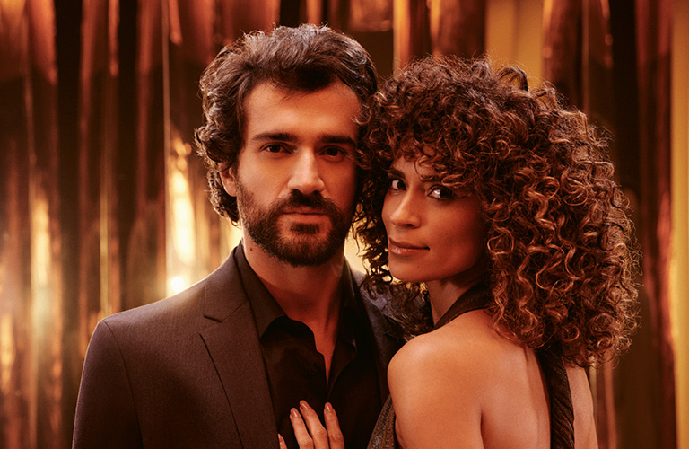 Foto de um casal em frente a cortinas marrons e douradas, com os rostos desfocados; a mulher encosta a mão no peito do homem, ambos vestidos de preto.