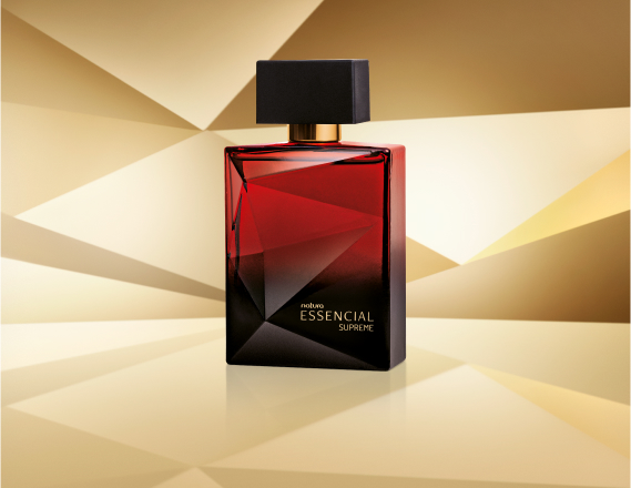 Frasco de Essencial Supreme Masculino, em tom de vermelho e preto e tampa preta, aparece iluminado sobre superfície e fundo dourados.