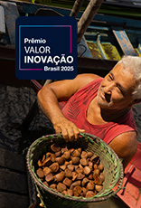 Um homem mais velho com cabelo grisalho está em um barco e carrega uma cesta cheia de castanhas do Brasil. No canto superior esquerdo, há um logotipo que diz "Prêmio VALOR INOVAÇÃO Brasil 2025".