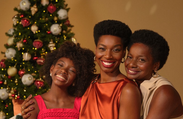 Mãe, filha e neta abraçadas e sentadas em um sofá, em frente à árvore de Natal enfeitada