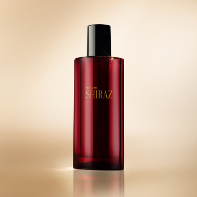 Desodorante Colônia Shiraz com frasco de vidro vermelho-escuro ao lado da caixa vermelha com detalhes em preto. Ambos estão sobre superfície reflexiva escura, com fundo em degradê de vermelho.