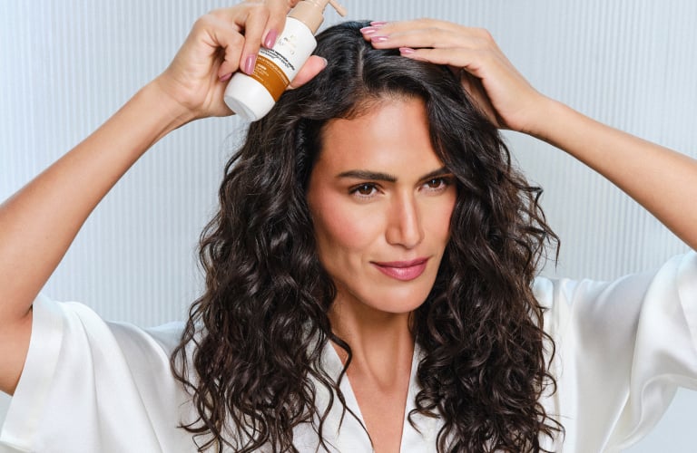 Mulher com cabelo longo ondulado aplicando tônico capilar Lumina