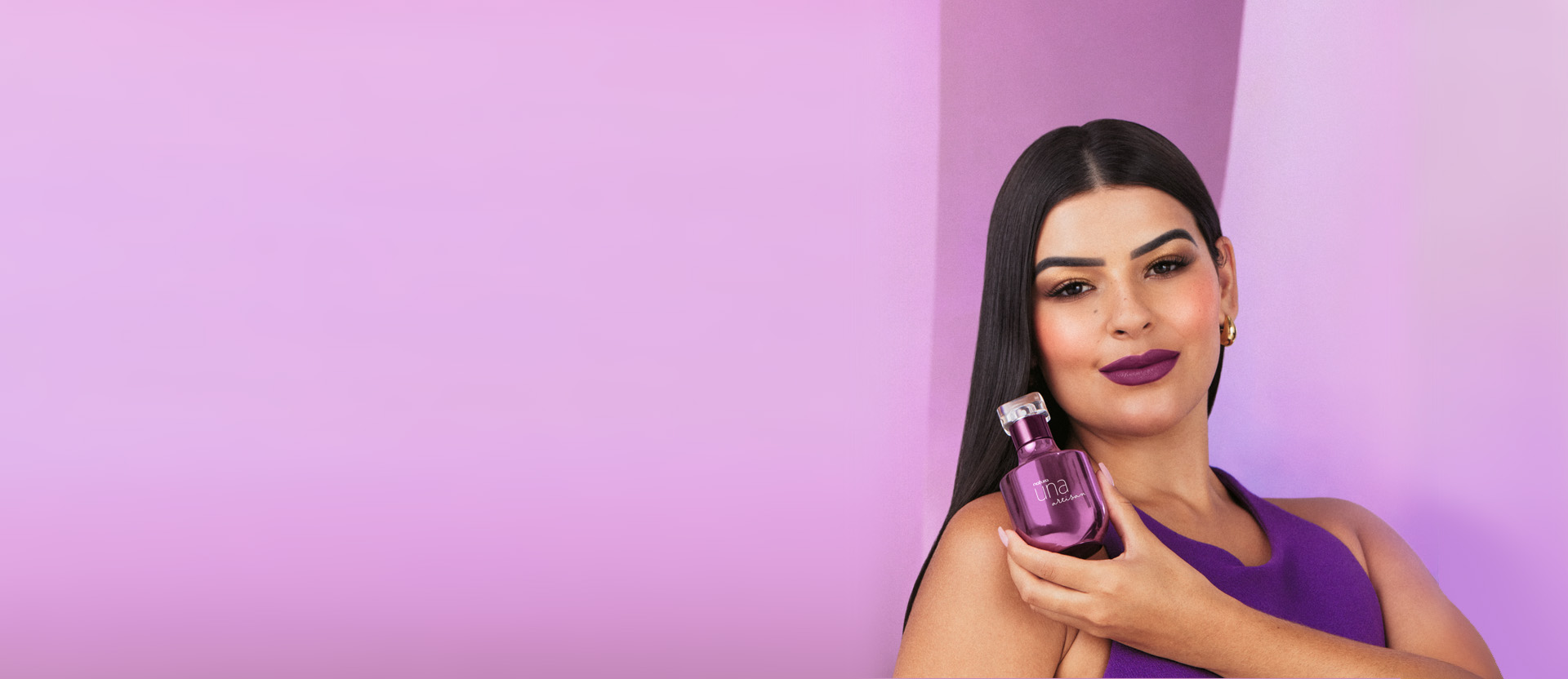 Foto de mulher de cabelos lisos e castanhos, segurando uma fragrância Natura Una, à direita, em fundo rosa.