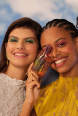 Duas mulheres olham para a câmera. À esquerda, mulher de pele clara e cabelos castanhos com blusa branca e, à direita, mulher de pele negra e tranças segura uma fragrância Natura Humor Envolve.