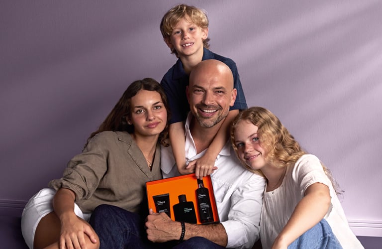 Foto com fundo neutro, mostrando pai sentado com caixa de presente aberta, cercado por três filhos ao lado e um menino sobre os ombros, em uma cena de família.