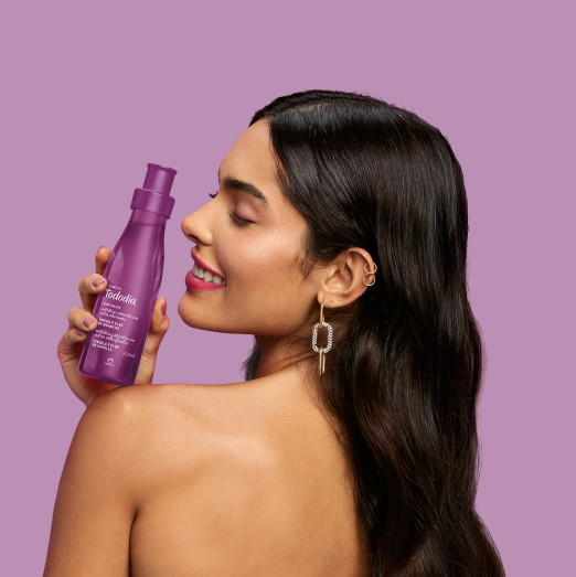 Mulher de cabelos longos segura um produto Natura Tododia sobre o ombro esquerdo, em fundo lilás.