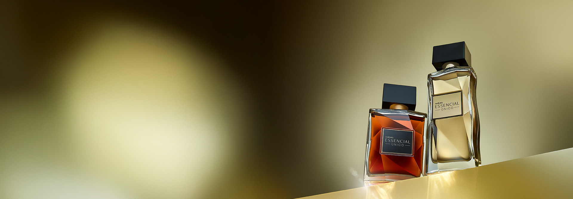 Essencial Único Masculino e Feminino, com frascos transparentes e tampas pretas, aparecem iluminados no canto direito da tela sobre superfície dourada e fundo em tons de preto e dourado.