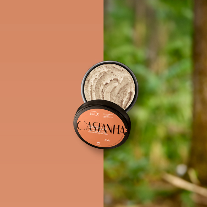 Pote aberto do Esfoliante Corporal Ekos Castanha, com textura cremosa bege, tampa marrom e rótulo laranja. Ao lado, castanhas cortadas sobre fundo marrom e verde desfocado.