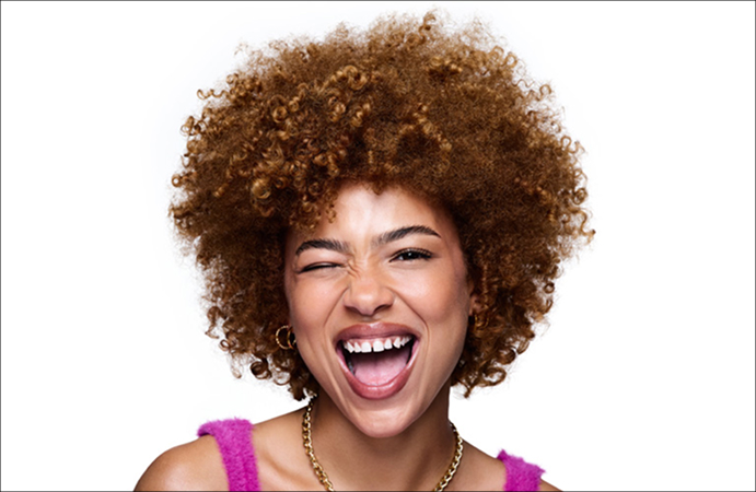Foto de uma mulher sorrindo com cabelos crespos volumosos, blusa rosa e colar dourado. Fundo branco.