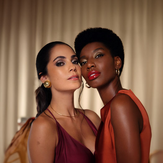 Mulheres com maquiagens marcantes: à esquerda, branca de cabelos castanhos presos, de vinho e brincos dourados, à direita, negra de cabelos curtos, de laranja e brincos de argola dourados, em fundo em tons de bege.