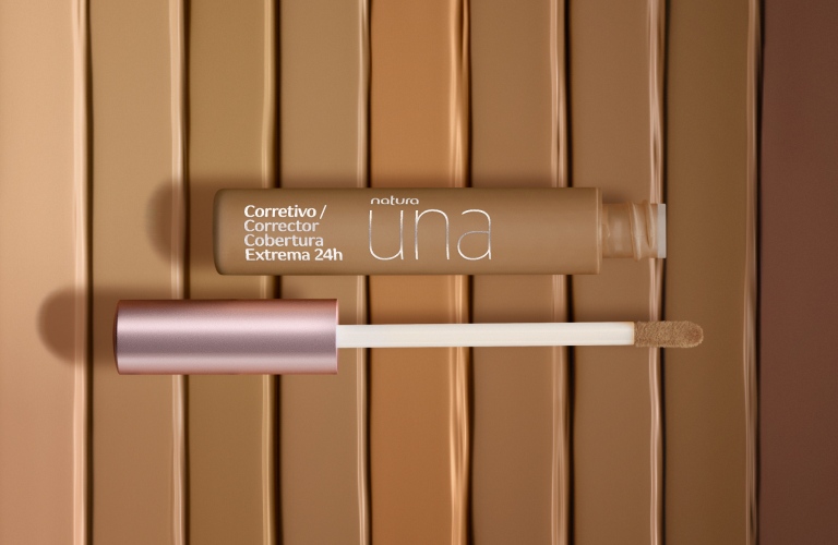 Corretivo Una posicionado na vertical com seu pincel ao lado, fora da embalagem. O fundo tem vários tons de marrom