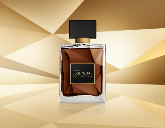 Frasco de Essencial Único Masculino, em vidro translúcido com fragrância em tons de âmbar e tampa preta, aparece iluminado sobre superfície e fundo dourados.