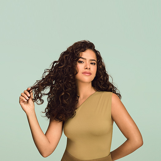Maisa segurando uma mecha de seu cabelo cacheado definido. Ela veste blusa bege sobre fundo verde-claro