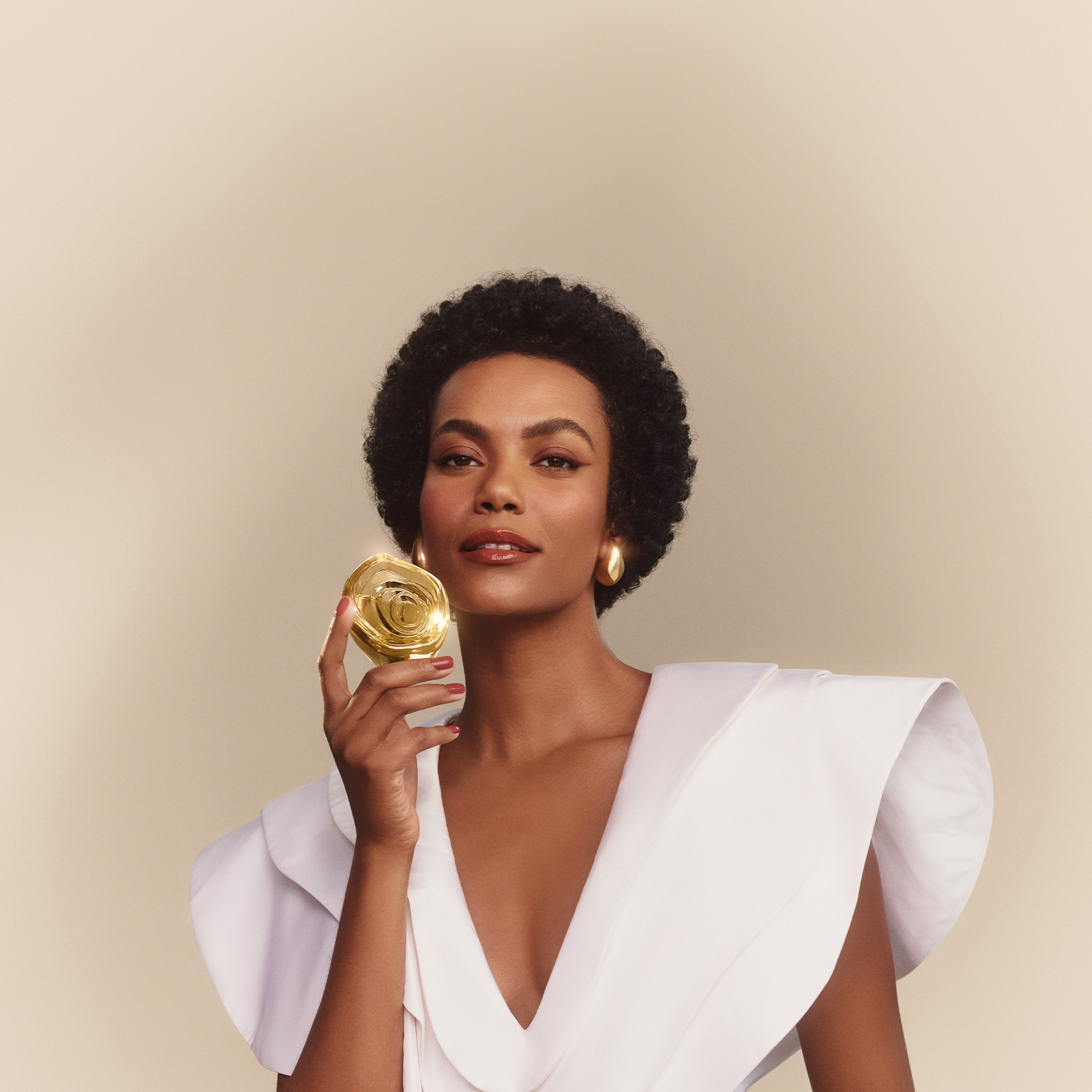 Mulher preta jovem de cabelo crespo curto segura o perfume Natura Aura Alba ao lado do rosto, com a parte em formato de rosa virada para frente. Ela veste branco e brincos dourados. O fundo é bege. 