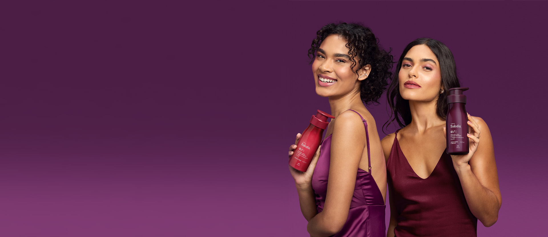 Duas mulheres, uma de cabelos cacheados e presos e outra de cabelos castanhos e soltos, seguram produtos Natura Tododia, em fundo vinho. Ambas usam vestidos de alças em tons de vinho.
