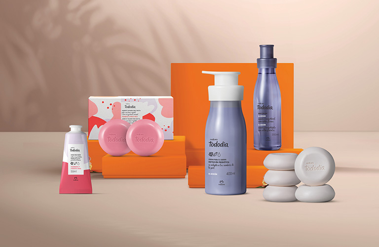 Foto de produtos de cuidado corporal sobre mesa bege, com fundo rosa claro e sombra de folhas; há frascos lilás, caixas, sabonetes e embalagens em rosa, laranja e branco.
