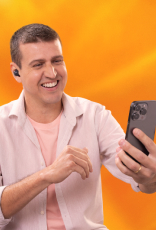 Imagem mostra homem de pele clara e cabelos curtos e castanhos vestindo camiseta rosada sobreposta por uma camisa branca. Ele usa fones de ouvido sem fio e sorri diante do celular. Fundo amarelo.