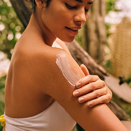 Mulher de pele morena, com cabelos castanhos presos, veste branco e aplica textura branca cremosa na pele do braço. Ela está em cenário de floresta em desfoque.