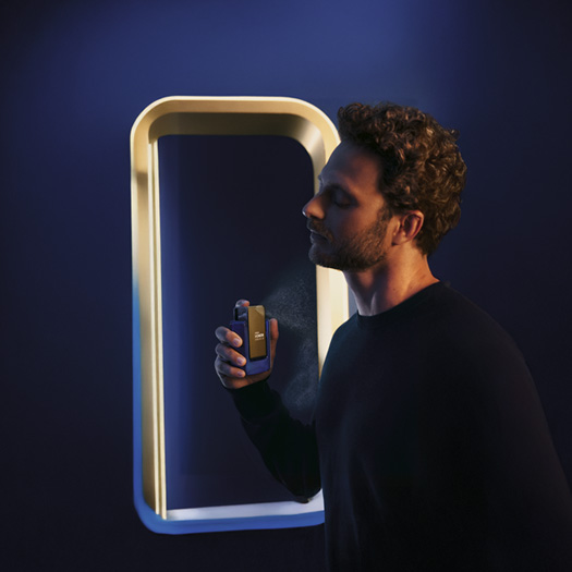 Homem de cabelos castanhos e cacheados aplicando no pescoço a fragrância Natura Homem Evolut.io, em fundo azul escuro e moldura branca iluminada.