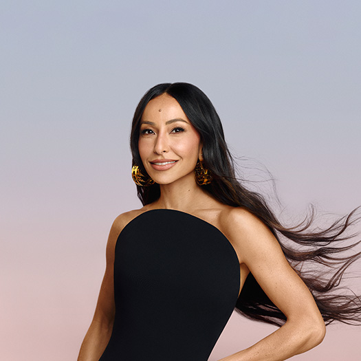 Fotografia de Sabrina Sato com cabelos soltos, usando brincos dourados e blusa preta, contra um fundo colorido suave.