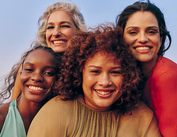Quatro mulheres sorrindo em um abraço coletivo. A mulher no canto superior esquerdo tem cabelos grisalhos longos. A mulher na parte inferior esquerda tem pele escura e cabelos trançados. A mulher no centro tem cabelos cacheados volumosos e castanho-avermelhados. A mulher no canto superior direito tem cabelos escuros e ondulados.