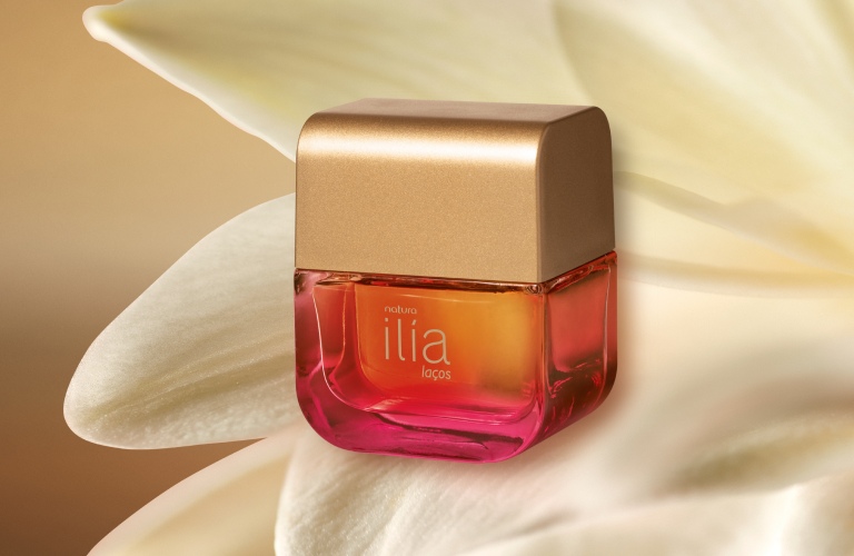 Perfume doce Ilía Laços posicionado na frente de uma flor branca. O fundo da imagem é dourado e o vidro do produto é rosa e laranja