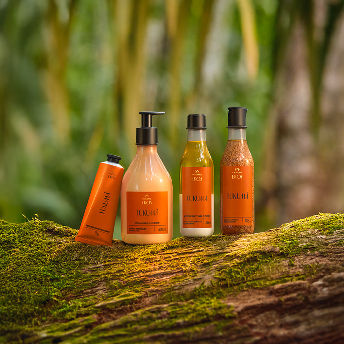 Kit Natura Ekos Tukumã com quatro produtos: bisnaga, frasco pump, frasco transparente e frasco com esfoliante, todos em tons de laranja sobre tronco coberto de musgo na floresta.