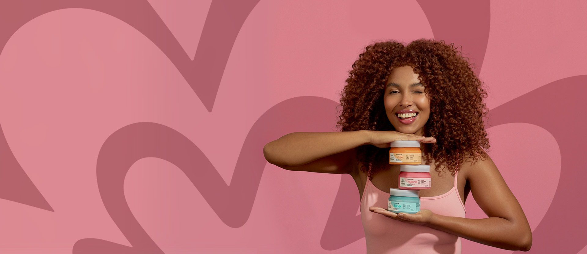 Mulher negra de cabelos cacheados e castanhos, sorrindo e segurando três potes de máscara de cronograma capilar Natura Tododia Cabelos nas cores laranja, rosa e azul-claro. Ela empilha os produtos em sua mão, com a máscara laranja no topo e a azul na base, próxima à sua mão. O fundo é rosa com um padrão abstrato da rosácea da Natura.