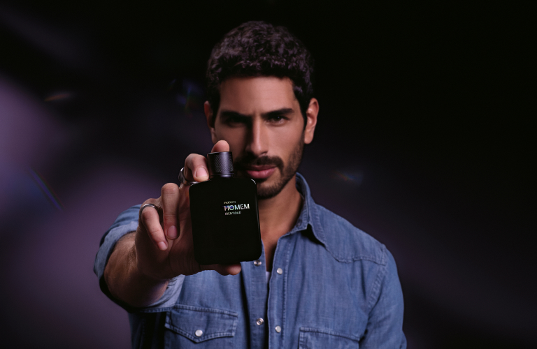 Foto com fundo escuro. Homem segura o frasco do perfume Natura Homem Identidad em direção à câmera.