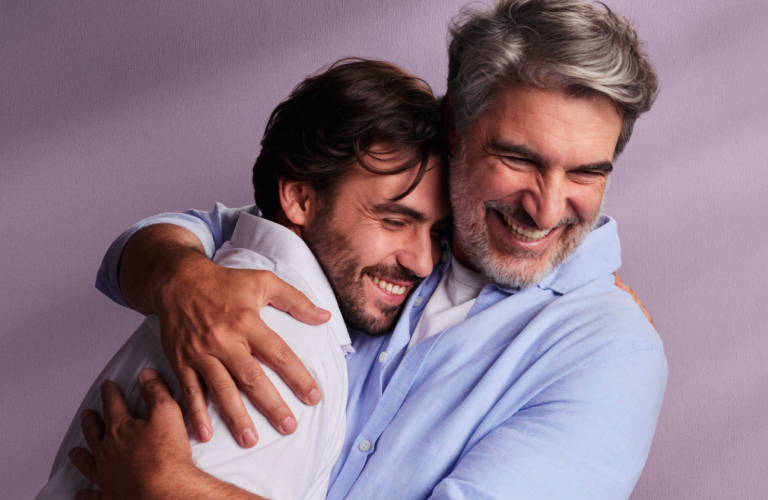 Foto de dois homens, um jovem e outro grisalho, abraçados sorrindo em um momento de carinho. Ao fundo, uma parede lilás com iluminação suave.