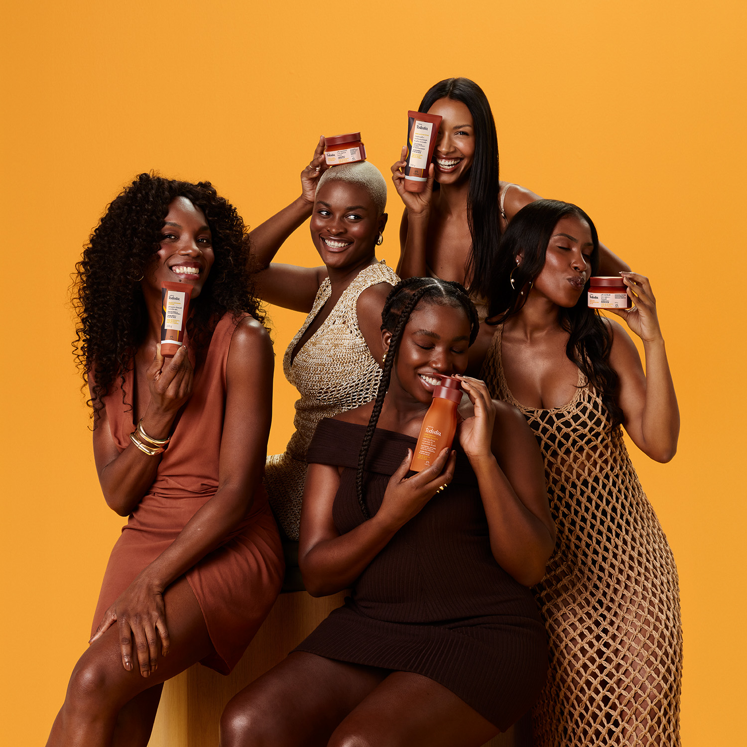 5 mulheres sorridentes usando vestidos em tons terrosos estão abraçadas lado a lado. Fundo amarelo vibrante.