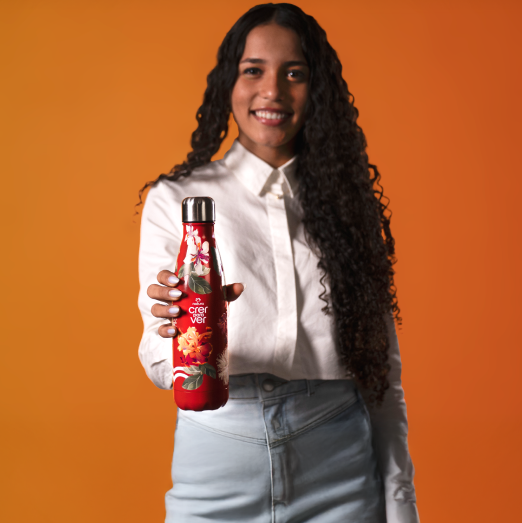Jovem de pele negra e cabelos compridos usa camisa branca e calça jeans e sorri. Ela segura a garrafa térmica Crer Para Ver, em tom de vermelho com estampa de flores e tampa metálica. Fundo laranja.