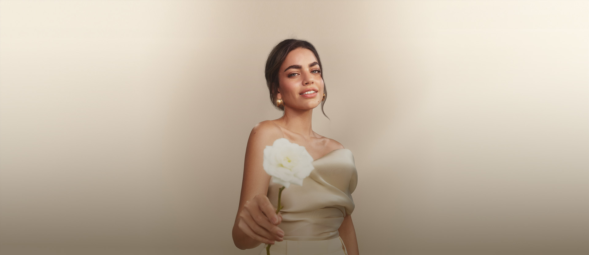 Foto de mulher com os cabelos presos segurando uma rosa branca, em fundo claro.