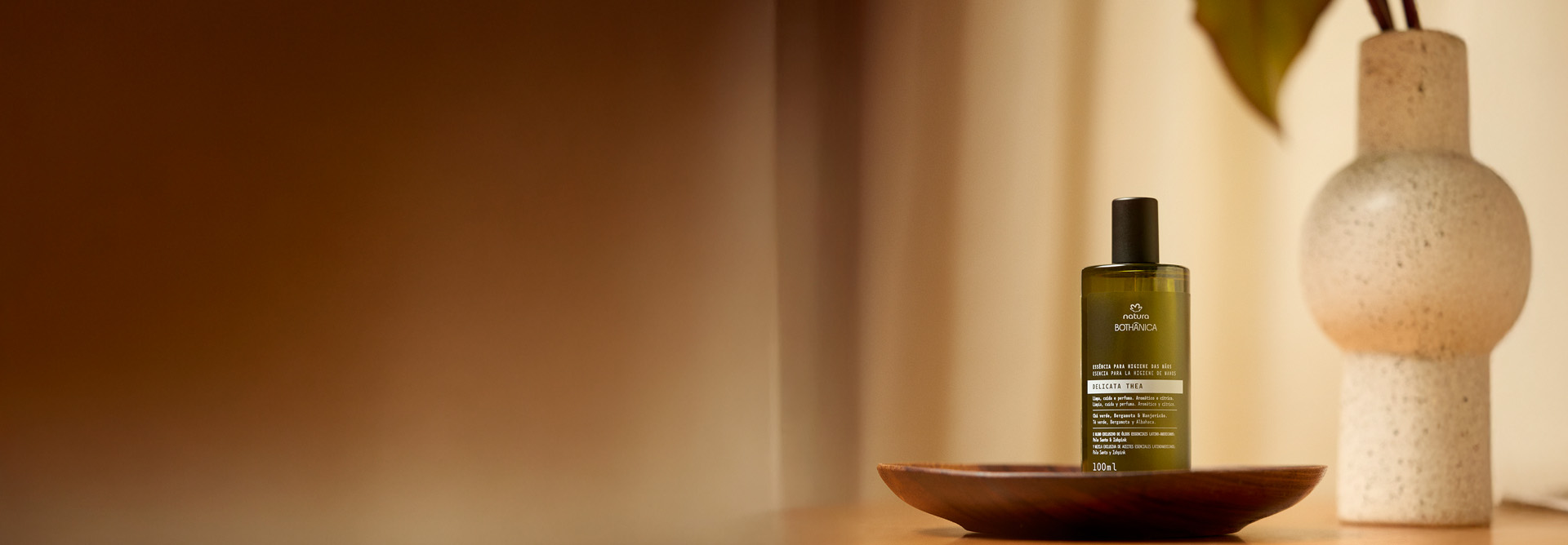 Essência para Higiene das Mãos Natura Bothânica Delicata Thea. Frasco verde escuro com detalhes brancos sobre prato de madeira escura em mesa clara, com vaso e planta ao fundo.